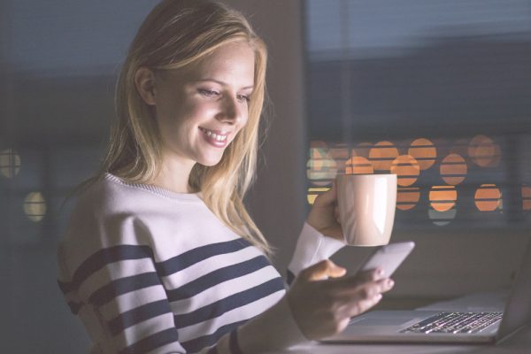Woman sat at desk with laptop on, smartphone in one hand, reading tips for becoming a self-employed blogger and a mug of tea in the other hand. It's night time and you can see a city scene out of the window.